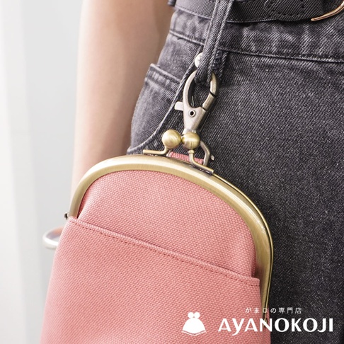 一個粉紅色帆布腰掛口金包，以古銅色金屬扣環及鏈條連接在深灰色牛仔褲的腰帶上，品牌標誌 AYANOKOJI 出現在圖片右下方。