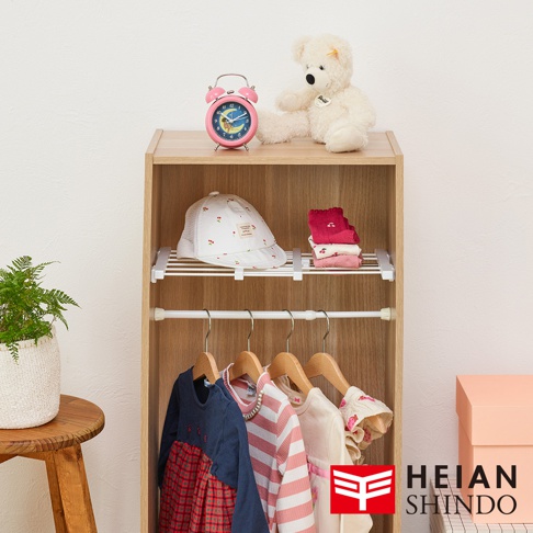 平安伸銅全功能伸縮縫隙置物架 SMR-24，展示架上擺放著可愛的粉紅色鬧鐘、白色櫻桃圖案的帽子、粉色疊放的襪子，以及可愛的白色泰迪熊。架子下方懸掛著幾件嬰幼兒衣物，包括一件深藍色和紅色格紋連身裙，以及粉白條紋上衣。旁邊還有粉色禮盒和綠色盆栽植物，下方角落印有 HEIAN SHINDO 的 Logo。