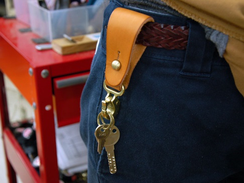淺棕色皮革製的直立型斜背包，透過銅製釦件固定於深藍色長褲的腰帶環上，並懸掛有兩支鑰匙。