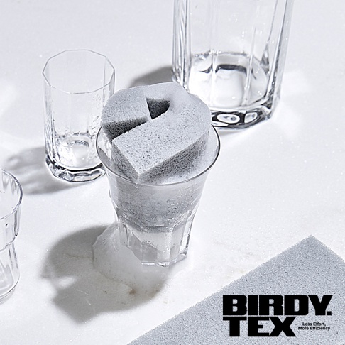 灰色耐用海綿刷置於充滿泡沫的玻璃杯內，旁邊還有其他透明玻璃杯，整體佈局乾淨明亮。