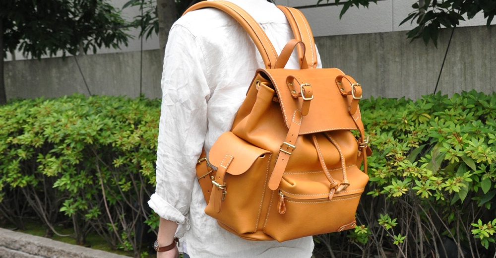 一個棕褐色皮革旅行後背包，有兩個前袋和束繩開口，配有搭扣和可調式肩帶。