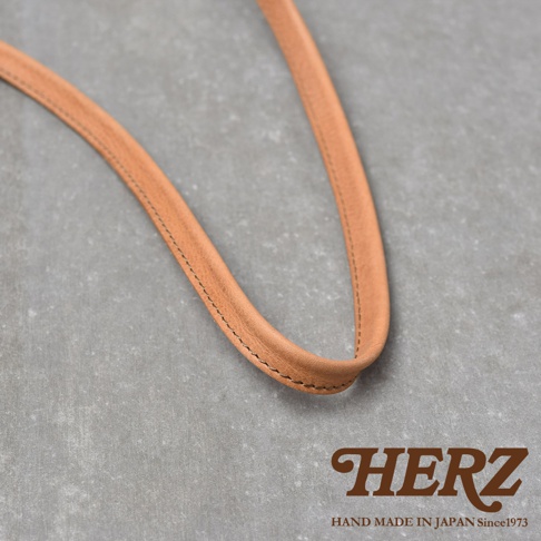 HERZ 品牌標誌，下方為 HAND MADE IN JAPAN Since1973 字樣，圖片主體為淺棕色皮革的斜背包帶，特寫鏡頭。