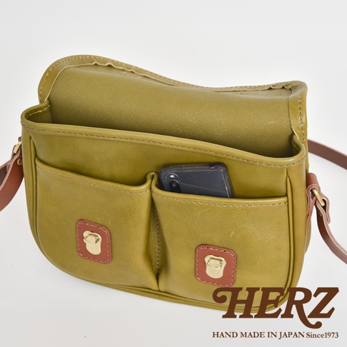 職人手作皮革傳統釣魚包，呈現橄欖綠色。包包為皮革材質，設有兩個前置口袋，其中一個口袋放入一支手機。包包帶有兩個金屬鎖扣，包包邊緣有縫線裝飾。