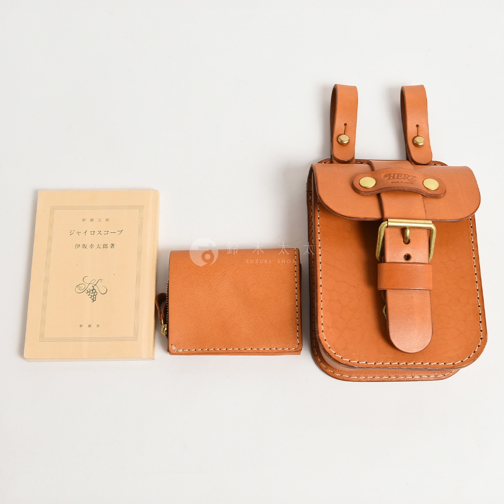 職人手作皮革經典腰袋，採用經典棕色牛皮製成，搭配黃銅釦帶與金屬卯釘。腰袋設計有兩條可懸掛的皮革帶，適合收納隨身物品。一旁另有同款皮革短夾和日文書籍。