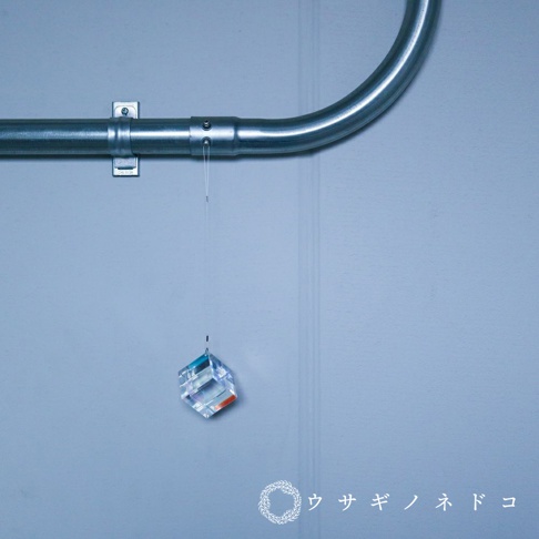 透明水晶立方體吊飾，底部有橙色和藍色光澤，懸掛於金屬管下方，背景為淺藍色牆面。