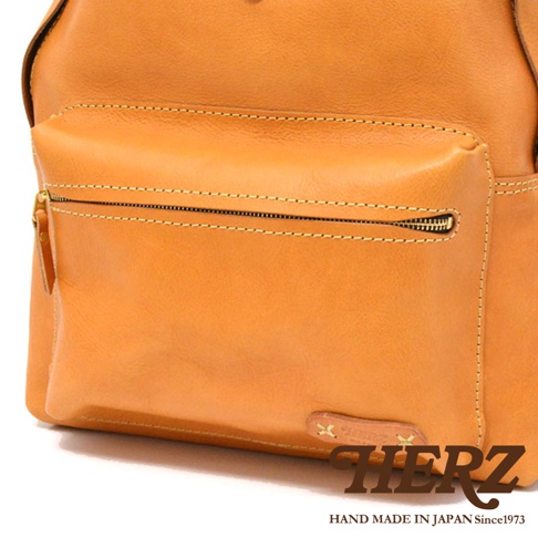 職人手作皮革休閒風後背包 R-93，採用天然皮革製成，顏色為溫暖的橘棕色。包身設計簡潔，前方有一個拉鍊口袋，車縫線明顯，展現精緻工藝。