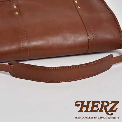 職人手作皮革復古郵差包，採用棕色皮革製作，細緻的車縫線與金屬铆釘裝飾，展現復古質感。包款結構簡潔，配有可調節的皮革背帶。