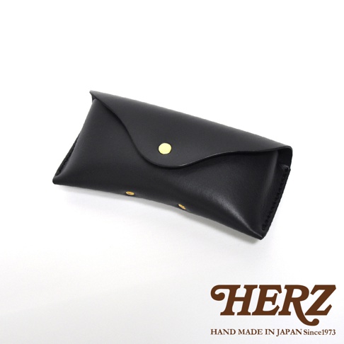 一個黑色皮革製成的眼鏡收納盒，盒蓋有金色按扣，底部和側面有縫線細節，右下角印有 HERZ HAND MADE IN JAPAN Since1973 字樣。