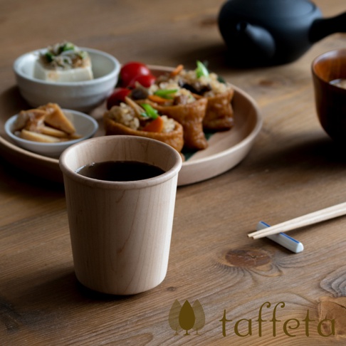 楓木寬口杯，杯身呈淺木色，材質為天然楓木，杯內盛有深色液體，置於木質桌面，背景為日式餐點。