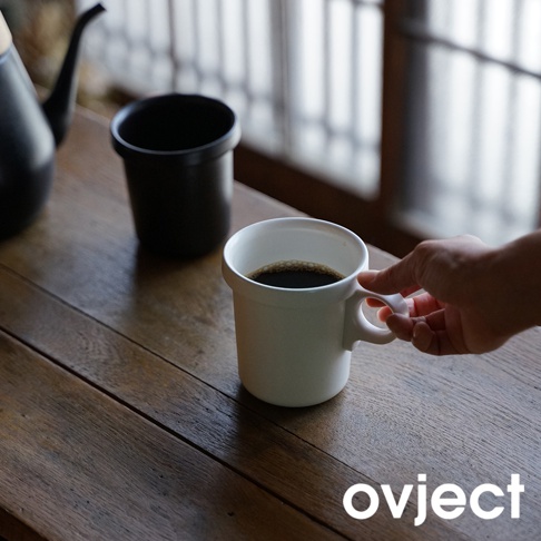 兩個簡約風格的琺瑯馬克杯，一個盛有咖啡，另一個為黑色空杯，擺放在木質桌面。