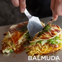 雙手使用兩支金屬鍋鏟，將加熱中的黑色電烤盤上的日式煎餅切開，煎餅內有麵條、蔬菜和醬料，表面撒有綠色粉末。