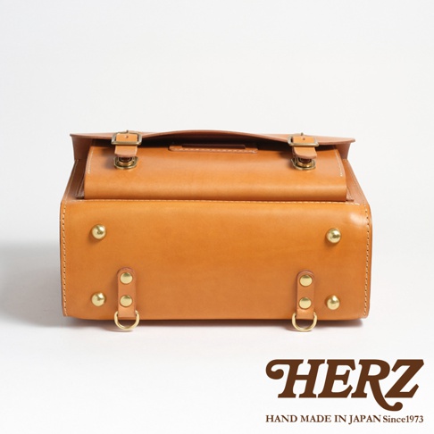 職人手作皮革橫式書包，淺棕色皮革，帶有金色釦環與铆釘，整體呈現復古風格，底部兩側有D型環設計。
