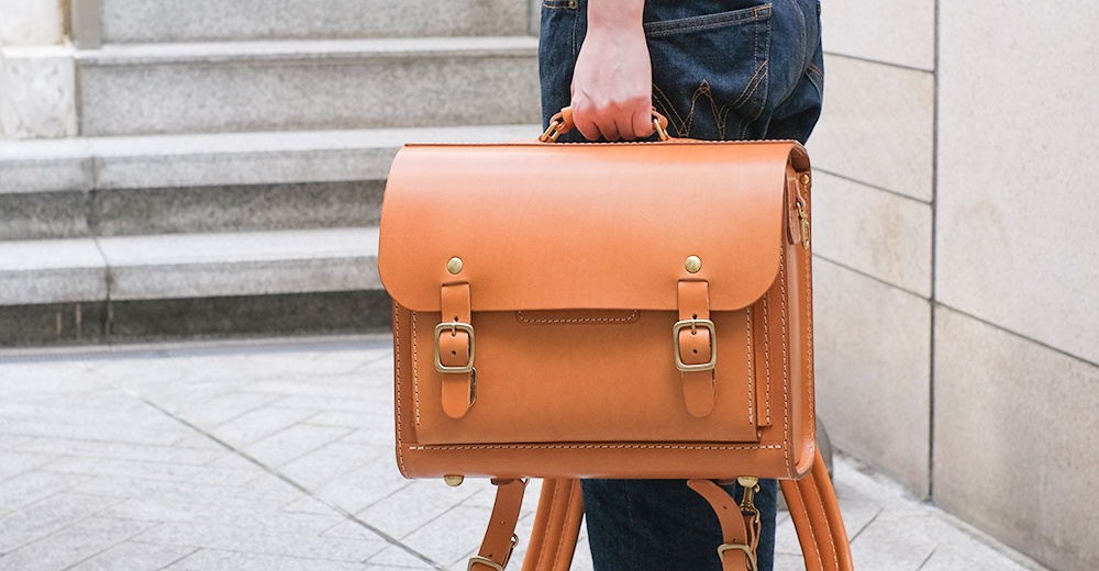 一個裝有皮帶和金屬扣的棕褐色皮革橫式書包，由一人手提著，背景是樓梯和牆壁。
