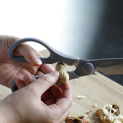 一手持用灰色廚房剪刀，另一手夾著花生，準備用剪刀將花生殼夾開，旁邊散落著花生殼。