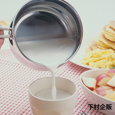 一只手握著不鏽鋼牛奶鍋，將白色液體（牛奶）倒入杯中。鍋子內部有刻度。背景有煎餅和炒蛋。