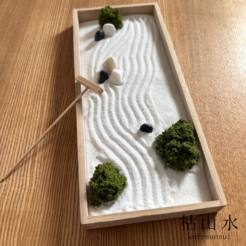 一個放置在木質桌面上的日本枯山水模型，長方形木盒內鋪滿白色細沙，沙面用小木耙繪製出層層波紋，並點綴著幾個綠色青苔塊、白色與黑色的石子，右下角印有黑色的枯山水日文與拼音字樣。