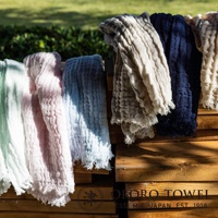 五條三層紗純棉掛脖毛巾，分別為淺綠、粉紅、淺藍、米白與深藍色，皆呈現皺褶紋理及流蘇邊緣，堆疊於木架上。