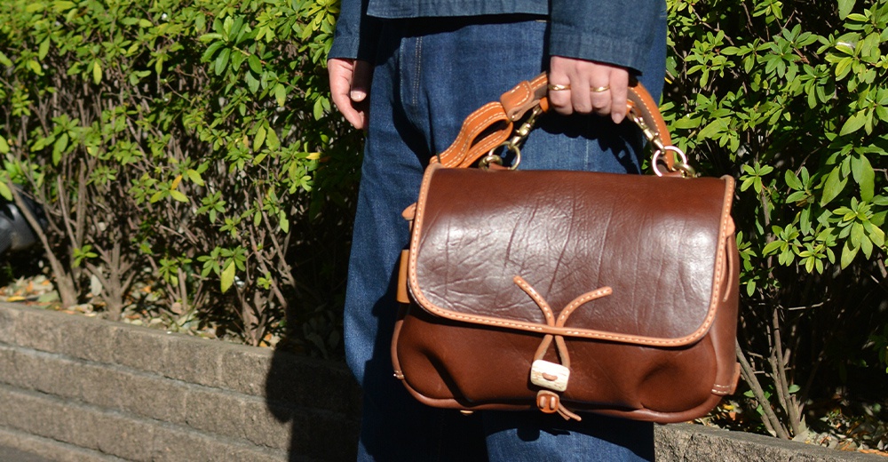 棕色皮革郵差包，配有鹿角造型的繩結閉合設計，可調節的皮革提把及肩帶，包身線條簡潔，質感厚實。