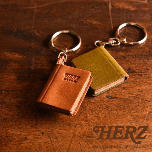 兩個皮革書本造型鑰匙圈，一個為棕色，印有「HERZ」及「MADE IN JAPAN」字樣；另一個為橄欖綠色。皆配有金色鑰匙圈。