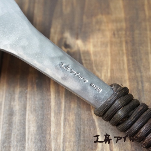 鋁製瀝水湯勺，勺柄處有錘目紋和「JAPAN」字樣，勺柄末端纏繞深褐色繩索，背景為木紋桌面。