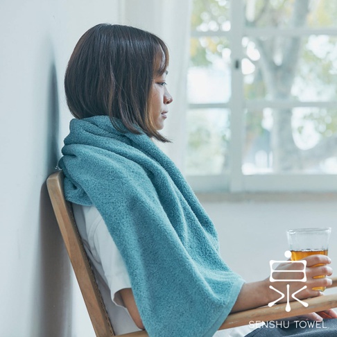 一位女性披著藍綠色長纖迷你浴巾，坐在木椅上，手中拿著一杯飲料。浴巾材質柔軟，質地明顯。