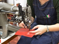 一台工業縫紉機正在縫製紅色皮革製品，一位身穿丹寧圍裙的女士正在操作。
