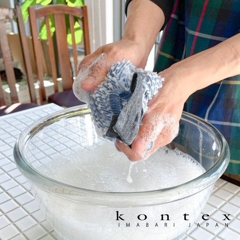 雙手正在搓洗一條藍白條紋的柔軟毛巾，毛巾中間夾著一塊灰色布料，旁邊是裝滿泡沫的透明玻璃碗。