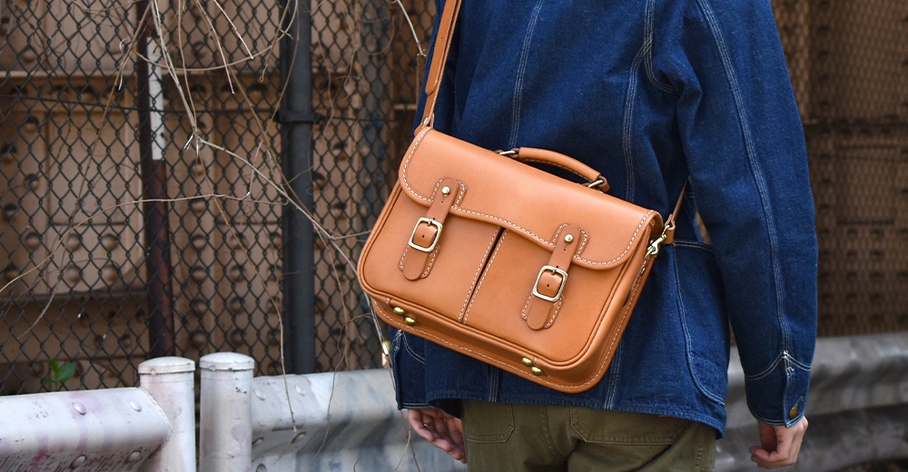 棕褐色皮革製成的手提斜背包，設有兩個皮帶釦和金色金屬扣環，車縫線明顯，可作為商務或休閒兩用包。