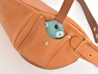 職人手作皮革斜背包，以天然淺棕色皮革製成，包身線條流暢，配有可調節的皮革肩帶和金屬扣環。包包側面有一個小口袋，露出部分印有圓形圖案的淺綠色內襯。