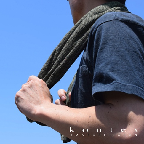 一名男子將一條深綠色、紋理明顯的長毛巾搭在肩上，背景為藍天。毛巾質地輕薄，觸感細膩，似乎由再生材料製成。