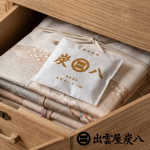 一個淺色的木質抽屜微微開啟，內部整齊地存放著摺疊好的和服或布料，布料上印有精緻的傳統紋樣。一個印有出雲屋炭八品牌標誌與文字的白色方形除濕木炭包放置在衣物上方，呈現出充滿日式生活氣息的居家收納場景。