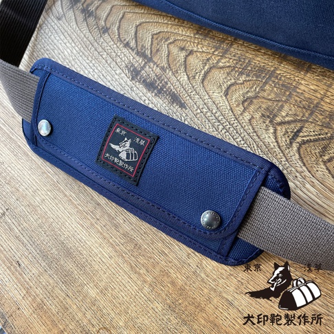 深藍色純棉帆布製成的肩墊，帶有織標和金屬扣，搭配灰色帆布背帶，放置於木紋背景上。