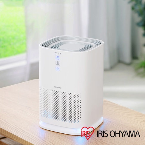 白色方形情境式空氣清淨機，置於木質桌面上。機身側面有許多小孔，頂部有風扇圖示、風速和電源按鈕。底部有藍色光暈，右下角是IRIS OHYAMA品牌標誌。