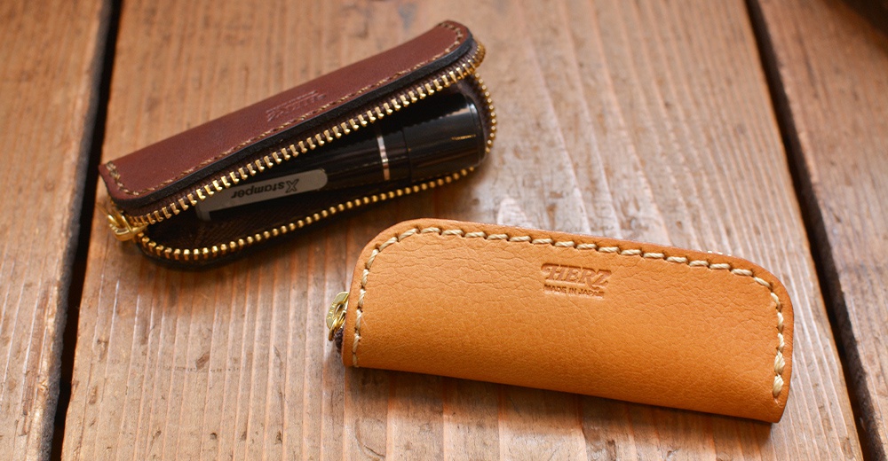 兩款職人手作皮革印章收納袋，一款為深棕色皮革，拉鍊半開露出內部印章；另一款為淺棕色皮革，表面印有品牌標誌，兩款皆有手工縫線與金色拉鍊。