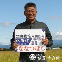 一位中年男性戴著眼鏡，身穿綠色外套，手持白色看板，上面寫著「契約栽培米 (輸出用) 北海道 稻穗の里協議 旭川市 - 高見 一典 ななつぼし」。背景是藍天白雲下的稻田和遠山。