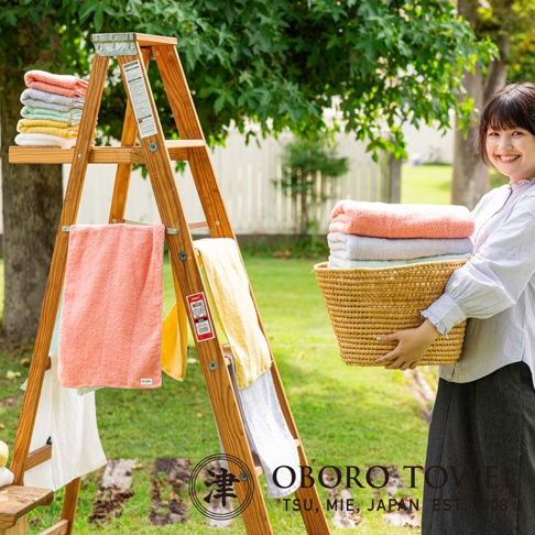 一疊粉橙色、淺藍色和淺綠色的毛巾放在一個編織籃子裡，由一位女士拿著。旁邊的木梯上掛著粉橙色、黃色和淺藍色的毛巾。