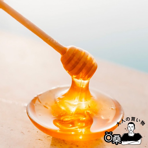 木製蜂蜜棒正在將金黃色的蜂蜜滴入一個淺碟中，上方印有「大人的購物」和一隻可愛的小熊圖案。