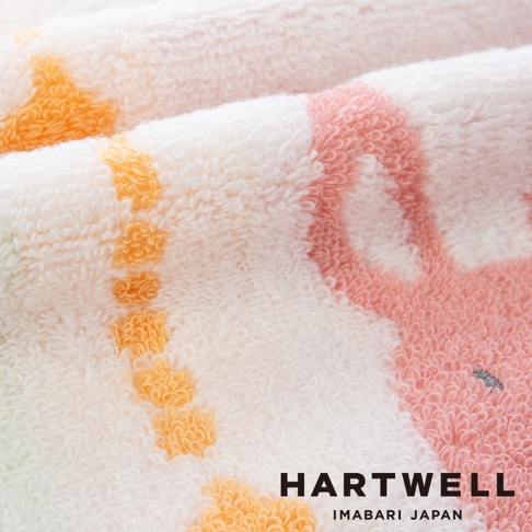 這是一張特寫鏡頭，呈現 Hartwell 今治森林動物刺繡手帕的柔軟毛圈布細節。淺色背景上編織著溫潤的粉色與明亮黃色圖案，纖維質感蓬鬆細緻，右下角處低調融入了 Hartwell Imabari Japan 的品牌標識。