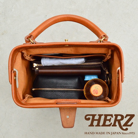 職人手作皮革Organ方形口金托特包，展現內部收納，包含黑色化妝包、咖啡色長夾、白色物品及圓形瓶裝物，皮革呈現自然的棕褐色。