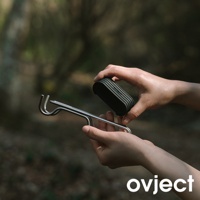 雙手各拿著一件產品：左手持金屬材質的彎鉤狀支架，右手持黑色、帶有條紋紋路的扁平小物，背景為模糊的綠色樹林。