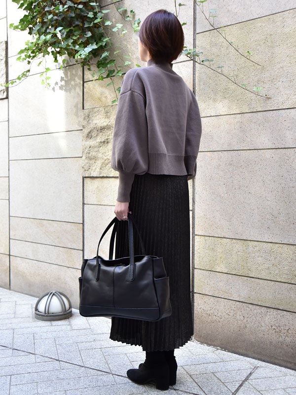 一位女士手提黑色皮革橫式肩背托特包，包包有兩個提把、正面有縫線設計，兩側有外袋。