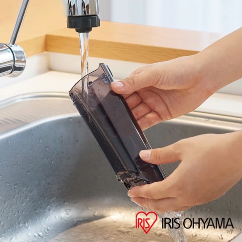 雙手正在水槽中沖洗攜帶式充電吸塵器 IC-H50 的透明深灰色集塵筒，集塵筒為塑膠材質，水龍頭的銀色金屬光澤清晰可見。