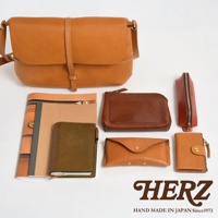 日本HERZ品牌手工製作的皮革產品，包含一個棕色斜背包、幾個小皮件如零錢包、卡夾，以及一本筆記本，背景為白色。