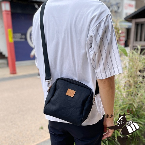 一個身穿白色條紋襯衫的人，斜挎一個黑色石洗帆布散步包，包上有一個淺棕色方形標籤。