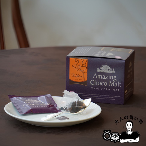 Lakshimi 品牌 Amazing Choco Malt 可可風味紅茶，茶包與一包裝產品放在白色盤子上，旁邊是產品盒裝，上方有日文標示「大人の買い物」及一位男性插畫。