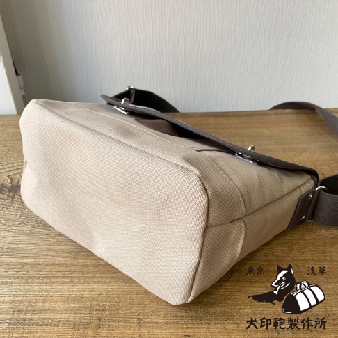 犬印純綿帆布雙扣郵差包，淺卡其色帆布材質，搭配深棕色皮革飾邊和肩帶，設有雙扣開合。