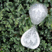 一個透明玻璃製成的泡泡沙漏，充滿細小的白色氣泡，置於綠色針葉植物背景中，沙漏呈現兩端膨大、中間收窄的沙漏形狀。