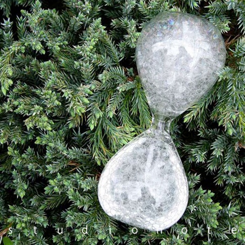 一個透明玻璃製成的泡泡沙漏，充滿細小的白色氣泡，置於綠色針葉植物背景中，沙漏呈現兩端膨大、中間收窄的沙漏形狀。