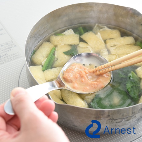 一只手拿著不鏽鋼瀝水湯匙，湯匙內盛有味噌湯料，湯匙上有兩根竹筷，湯匙正從裝滿油豆腐和青菜的鍋中撈起湯料。