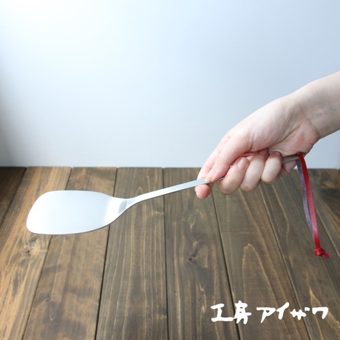 一只手正握著一支銀色不鏽鋼萬用鍋鏟，鍋鏟的鏟面為寬闊的橢圓形，手柄細長，末端綁著一條紅色的細繩。